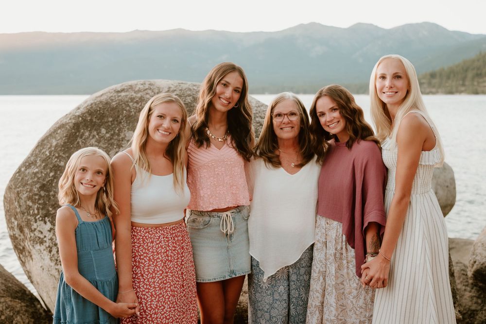 Six women and girls smiling together outdoors near a lake with mountains and large boulders in the background.