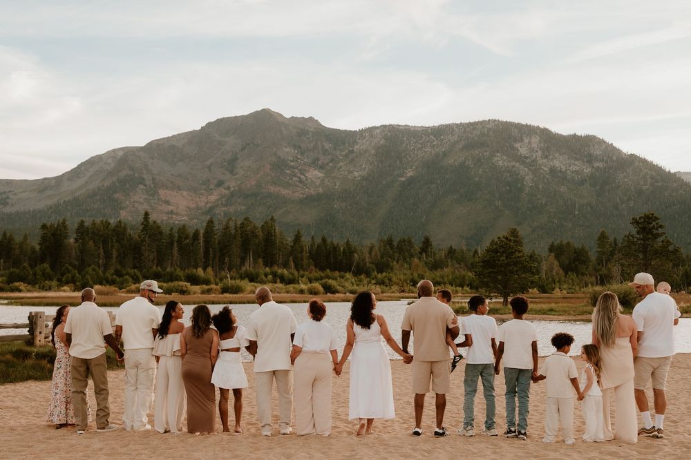 Large family group standing together on a sandy beach with a mountain backdrop and pine trees in neutral white outfits.