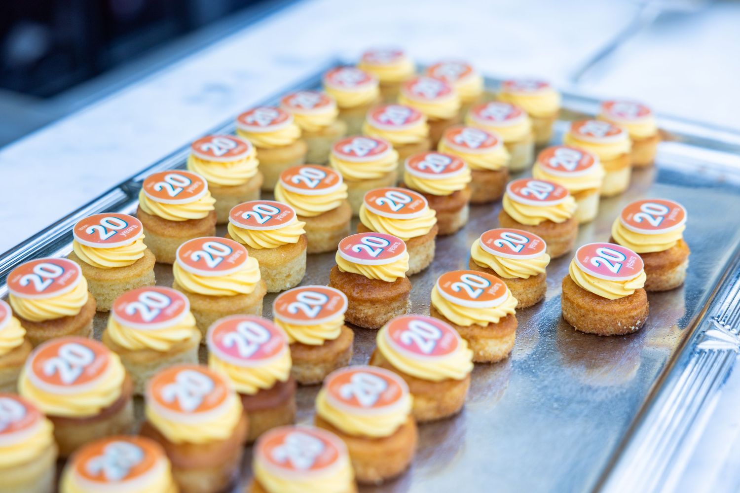Rows of decorated anniversary cupcakes with yellow frosting on a silver tray.