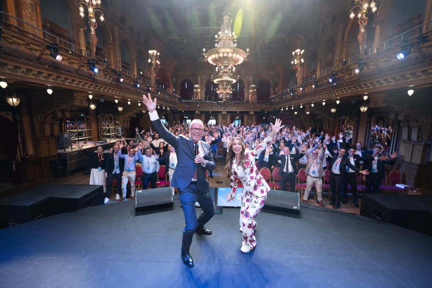 Two presenters on stage raising their arms to a cheering crowd in a grand ballroom.