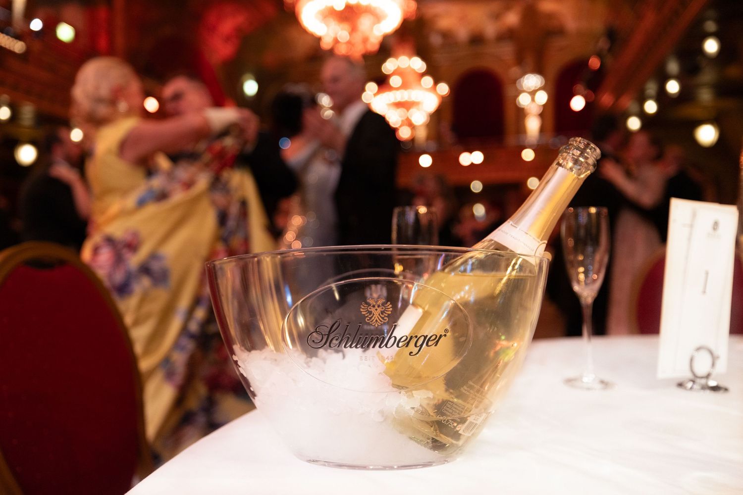 Champagne bottle chilling in ice bucket at elegant gala event with couples dancing in background.