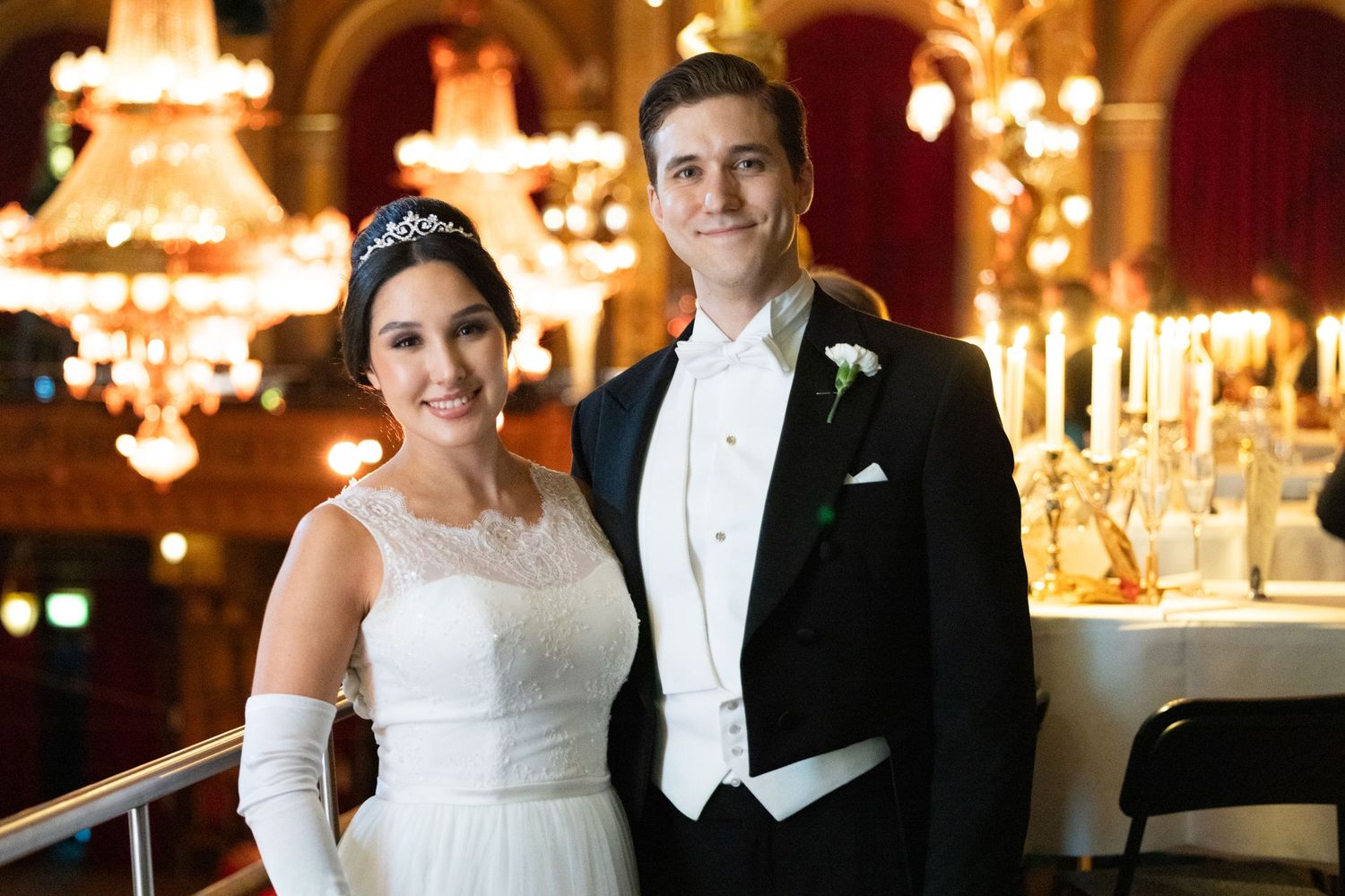 Elegant couple in formal attire posing at luxurious ballroom wedding reception with chandeliers.
