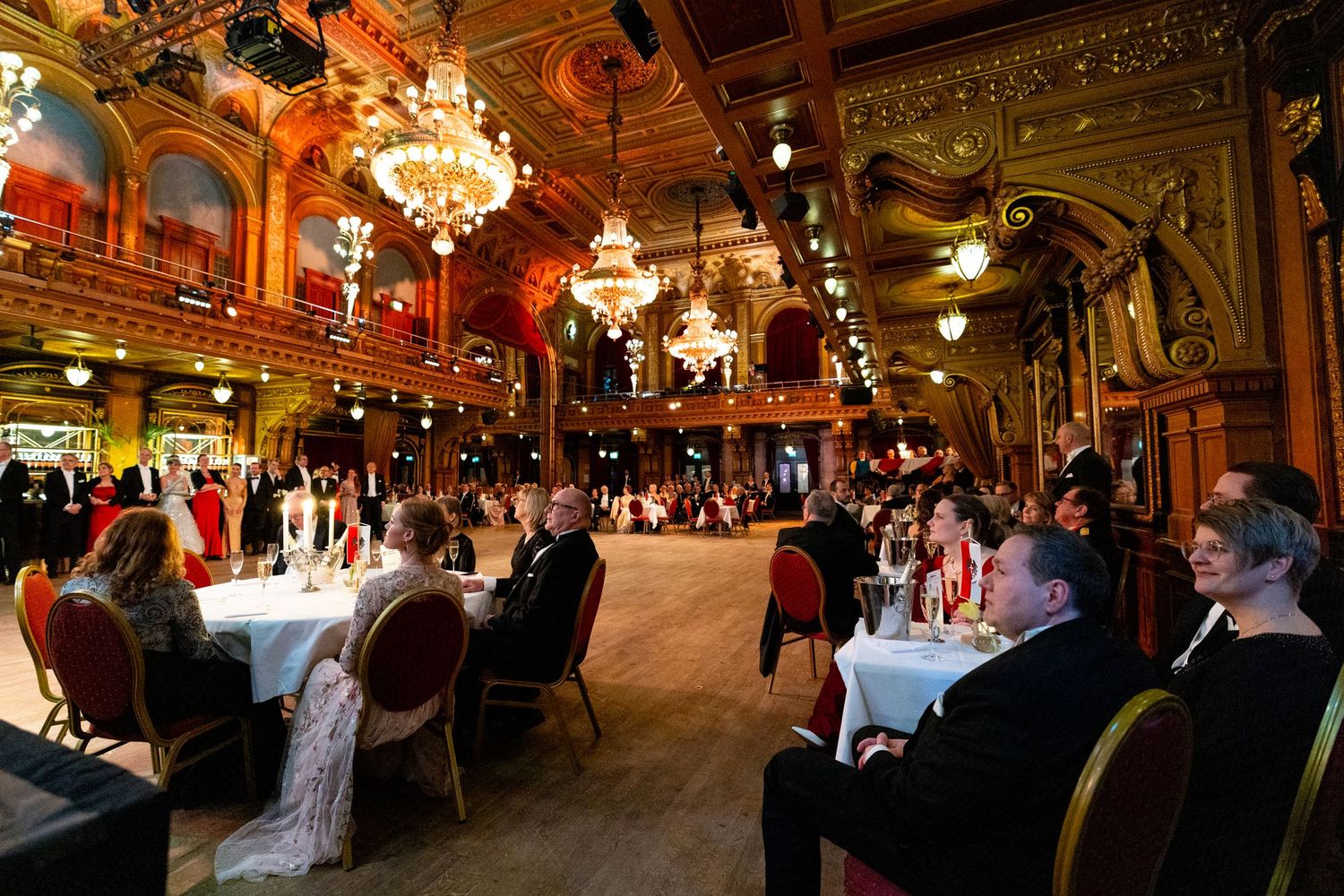 Elegant gala dinner in a grand ballroom with chandeliers, guests seated at formal tables.