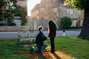 Man proposing on one knee to woman on grass near bench, red rose petals scattered around, charming stone building backdrop in Broadway.