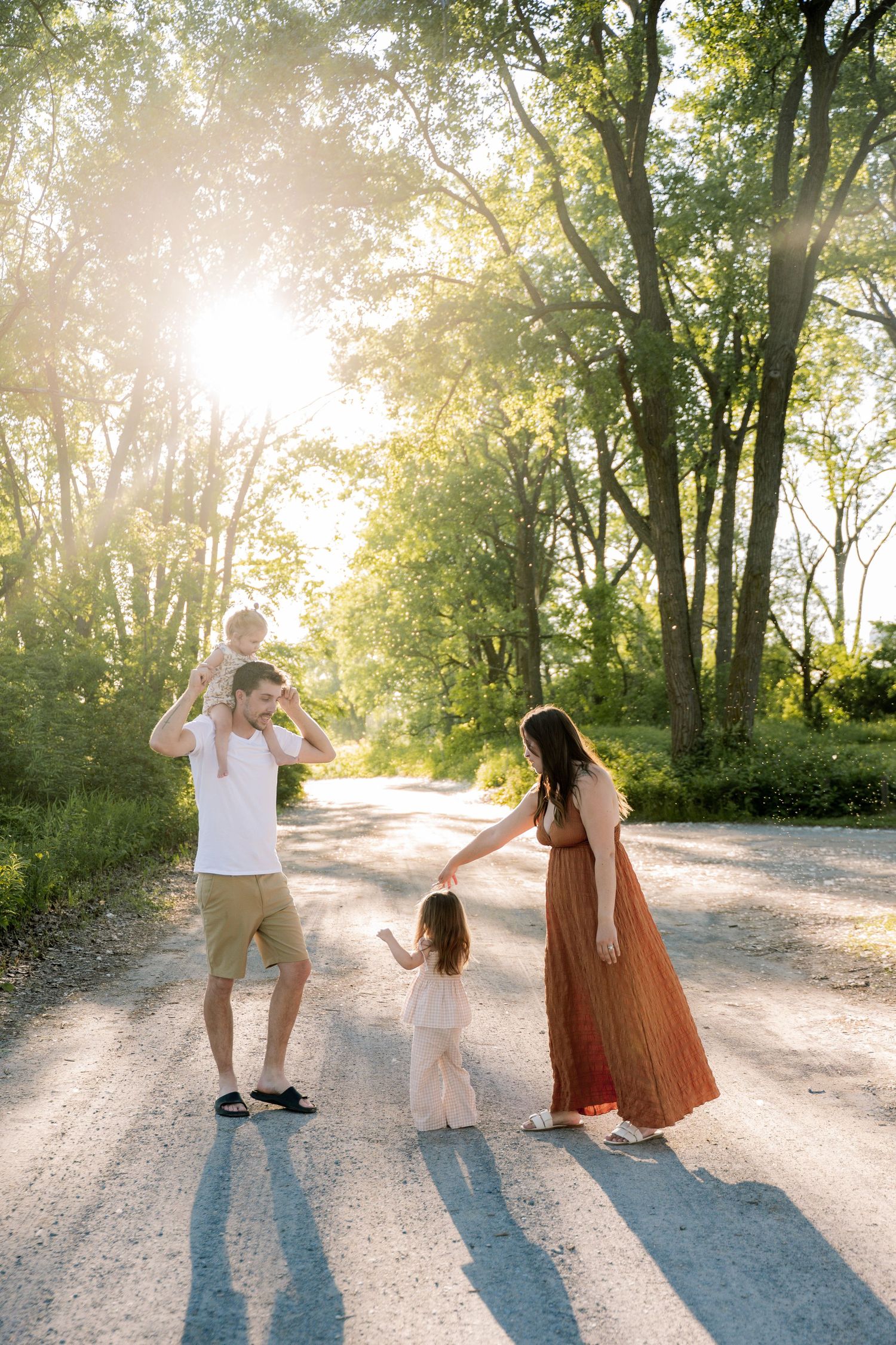 Family of four enjoying a sunny walk on a tree-lined gravel road, with golden backlight and children playing.