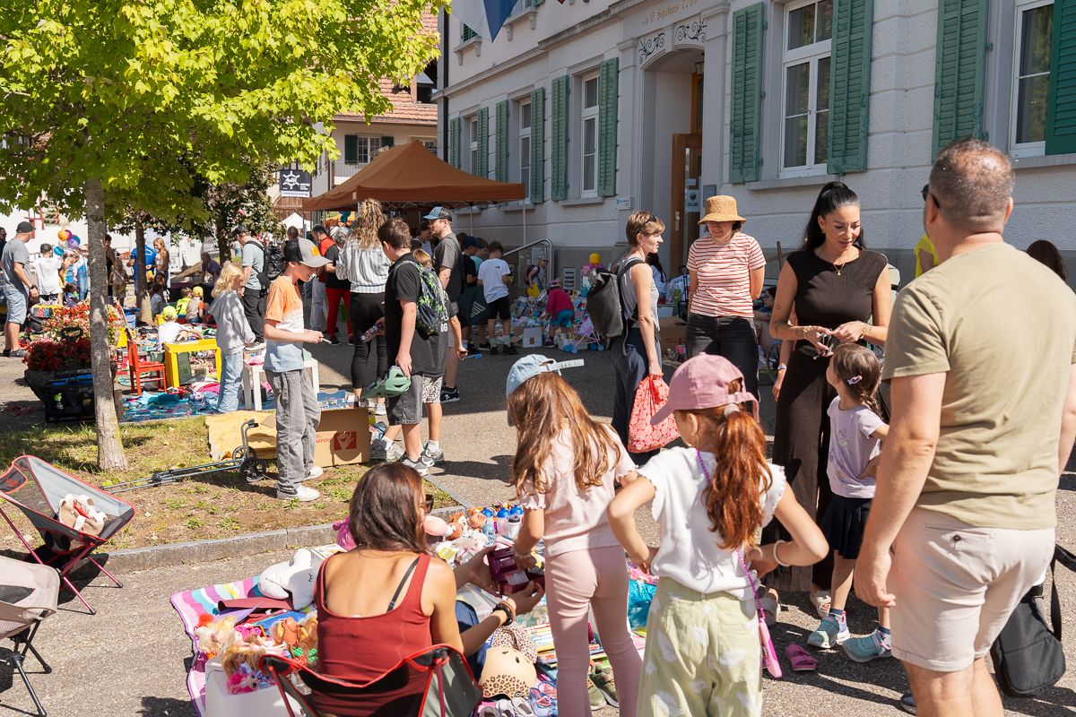 Families browse colorful items at an outdoor flea market on a sunny day in a European town square.