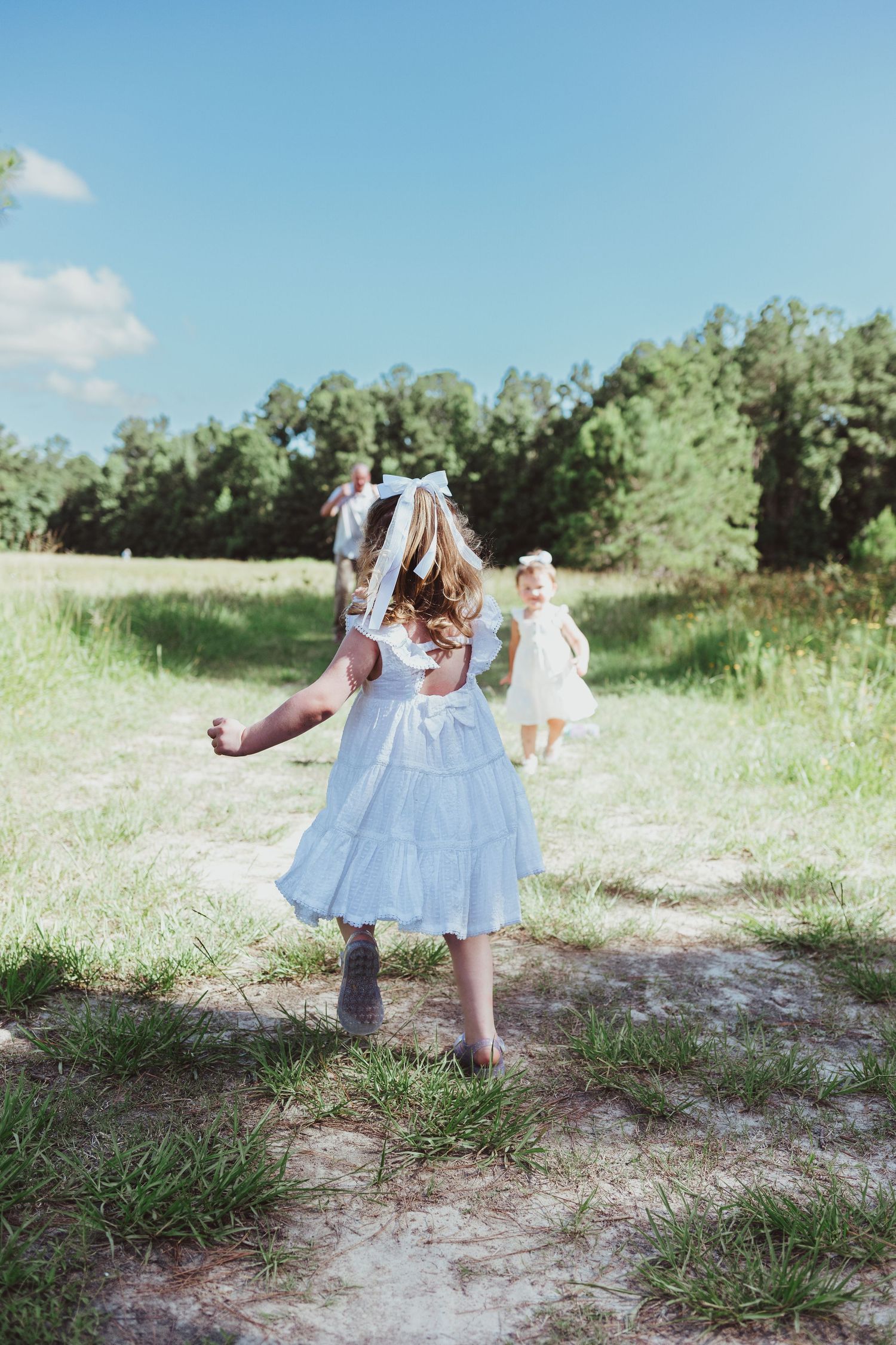Girls in white and blue dresses playing in a sunny green meadow with trees in the background.