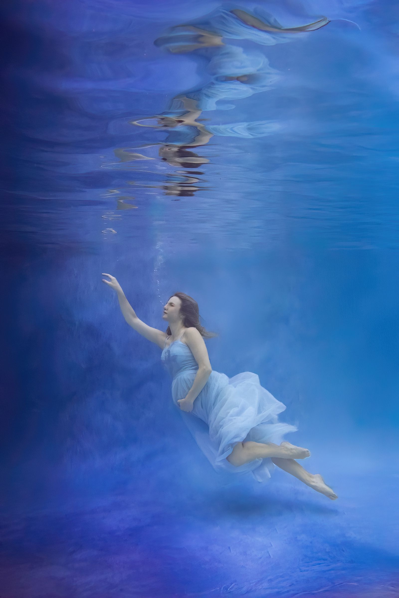 Expectant mother floating in a flowing blue gown beneath a mirrored reflection.