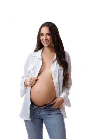 Expectant mother in jeans and an open white shirt smiling against a bright background.
