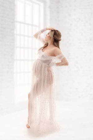 Expectant mother in a sheer white gown standing in front of a bright studio window.