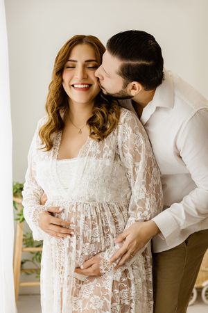Expectant couple smiling together in light neutral clothing beside a bright window.