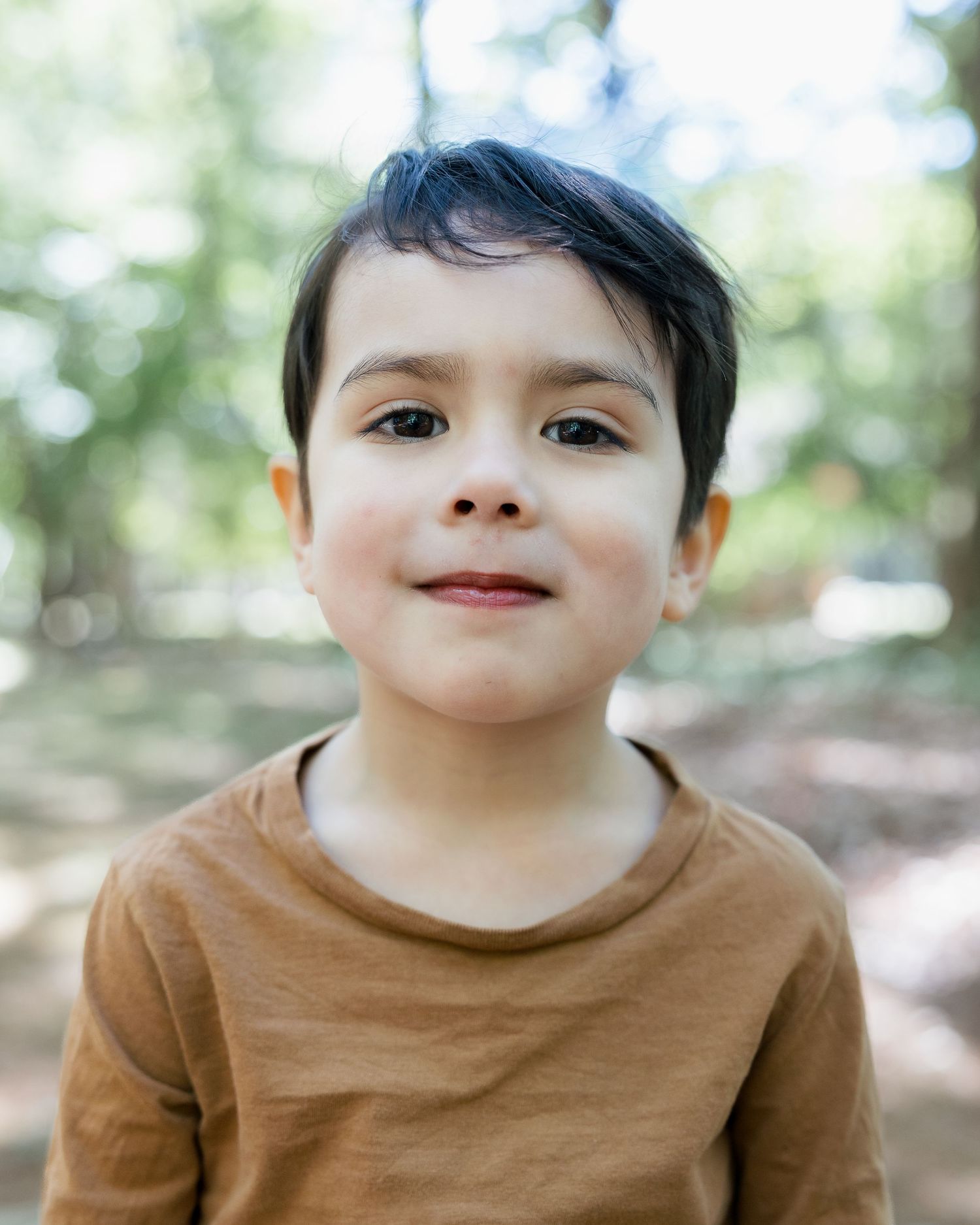 Young boy with dark hair wearing a brown long-sleeve shirt smiling outdoors in a park setting.