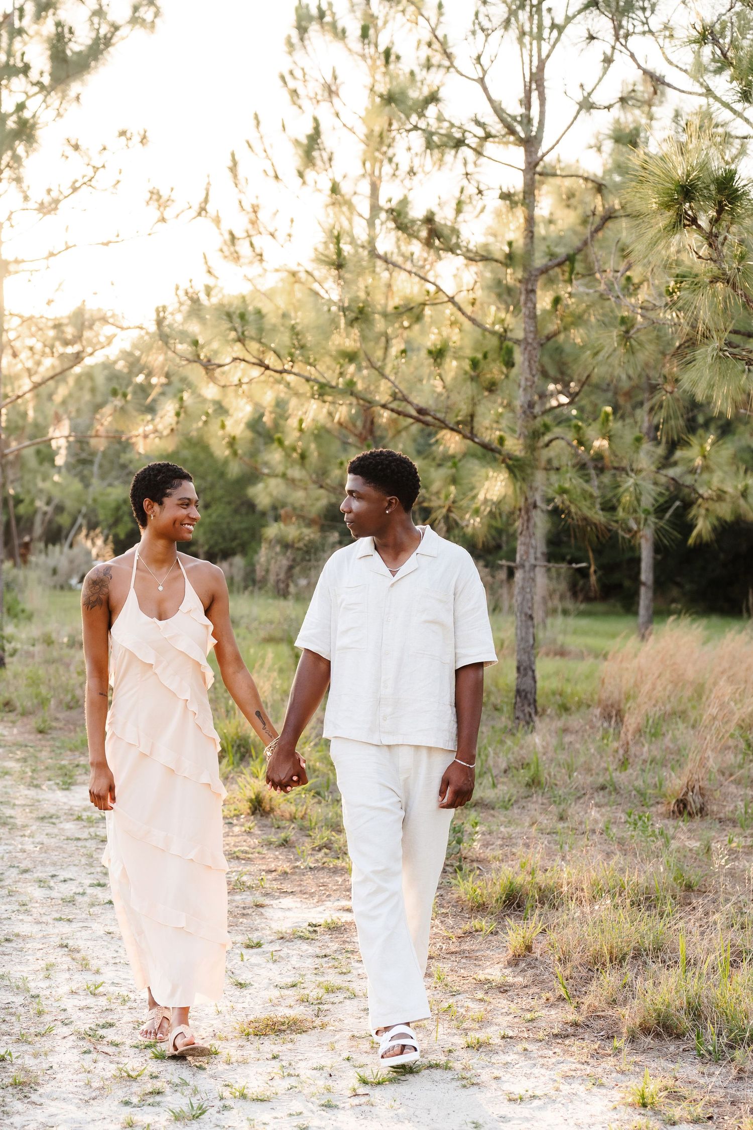 A couple in white outfits holds hands while walking on a sandy path surrounded by pine trees in golden hour light.