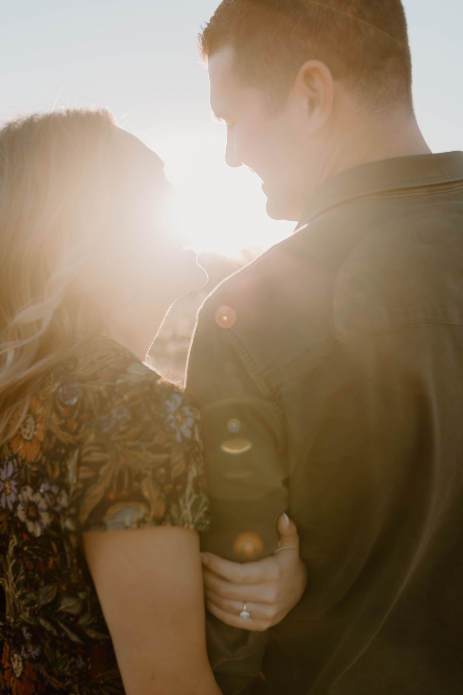 An engaged couple embraces in a golden sunset backlit glow, showcasing an engagement ring.