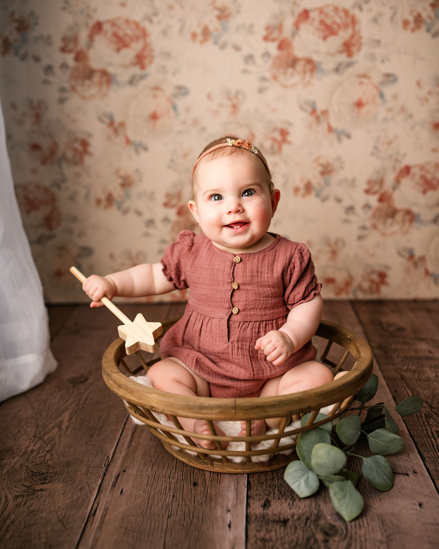 Smiling baby girl in rust romper sitting in wicker basket holding wooden star wand.