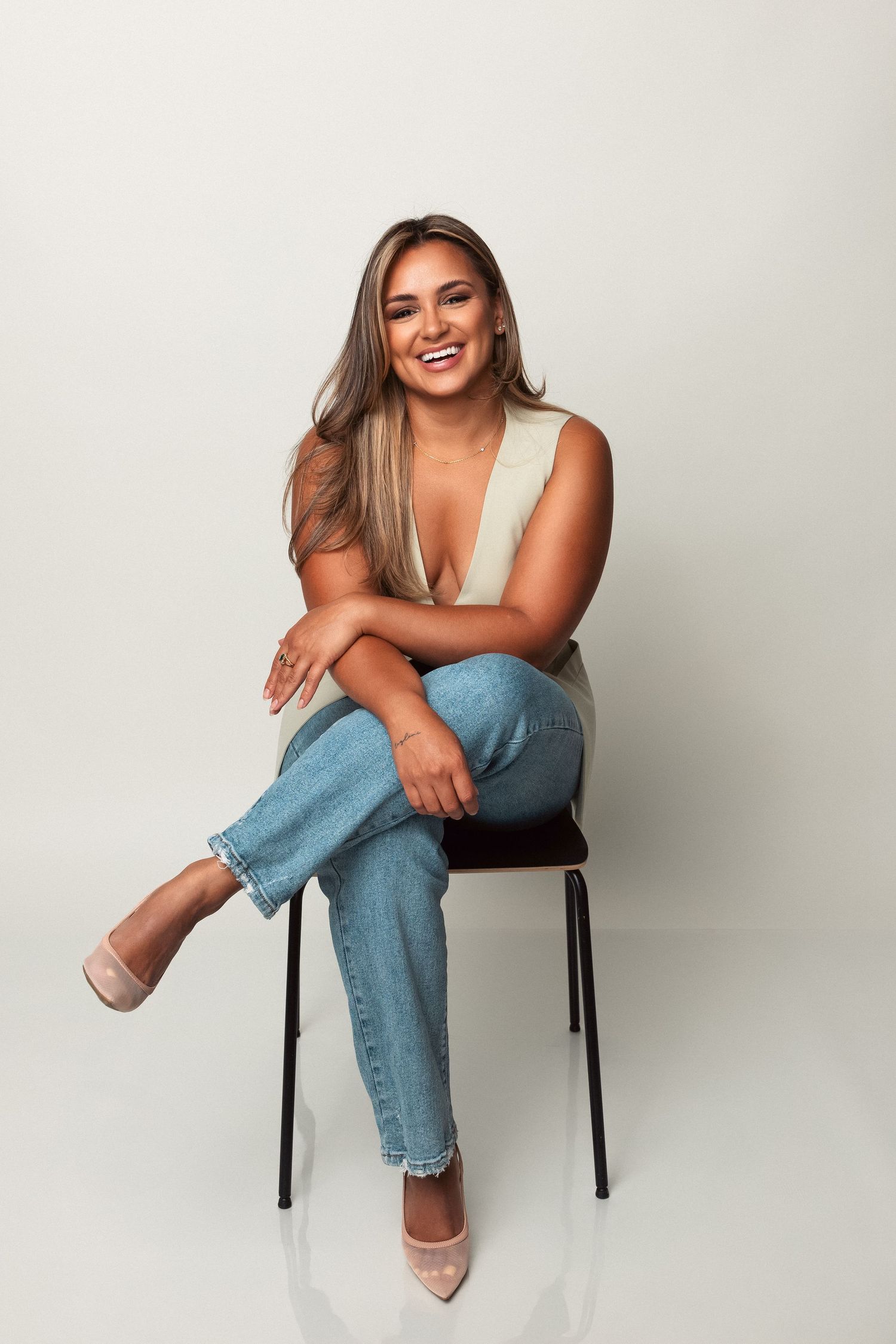 A woman in light colored top and jeans sits casually on a chair against a white background, smiling warmly.