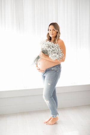 Maternity portrait of woman in jeans and floral top against white backdrop.