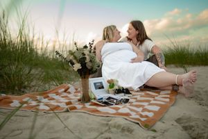 Two women share a romantic moment on a beach blanket at sunset, surrounded by flowers and photos.