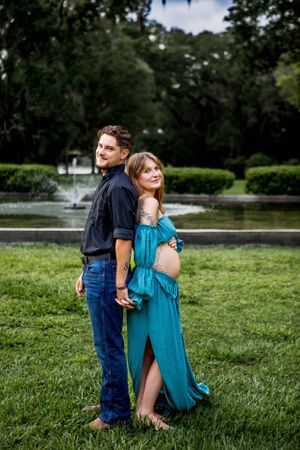 Couple poses back-to-back in park setting with fountain in background.