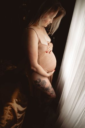 Artistic maternity photo silhouetted against window light shows pregnant figure with arm tattoo in intimate boudoir setting.
