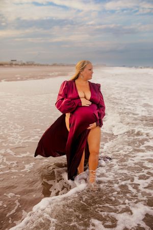 Figure in red dress poses dramatically in ocean waves at beach during sunset as surf splashes around them.