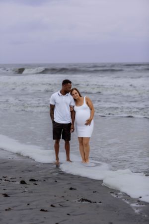 Couple walking along beach shoreline at sunset wearing white clothing.
