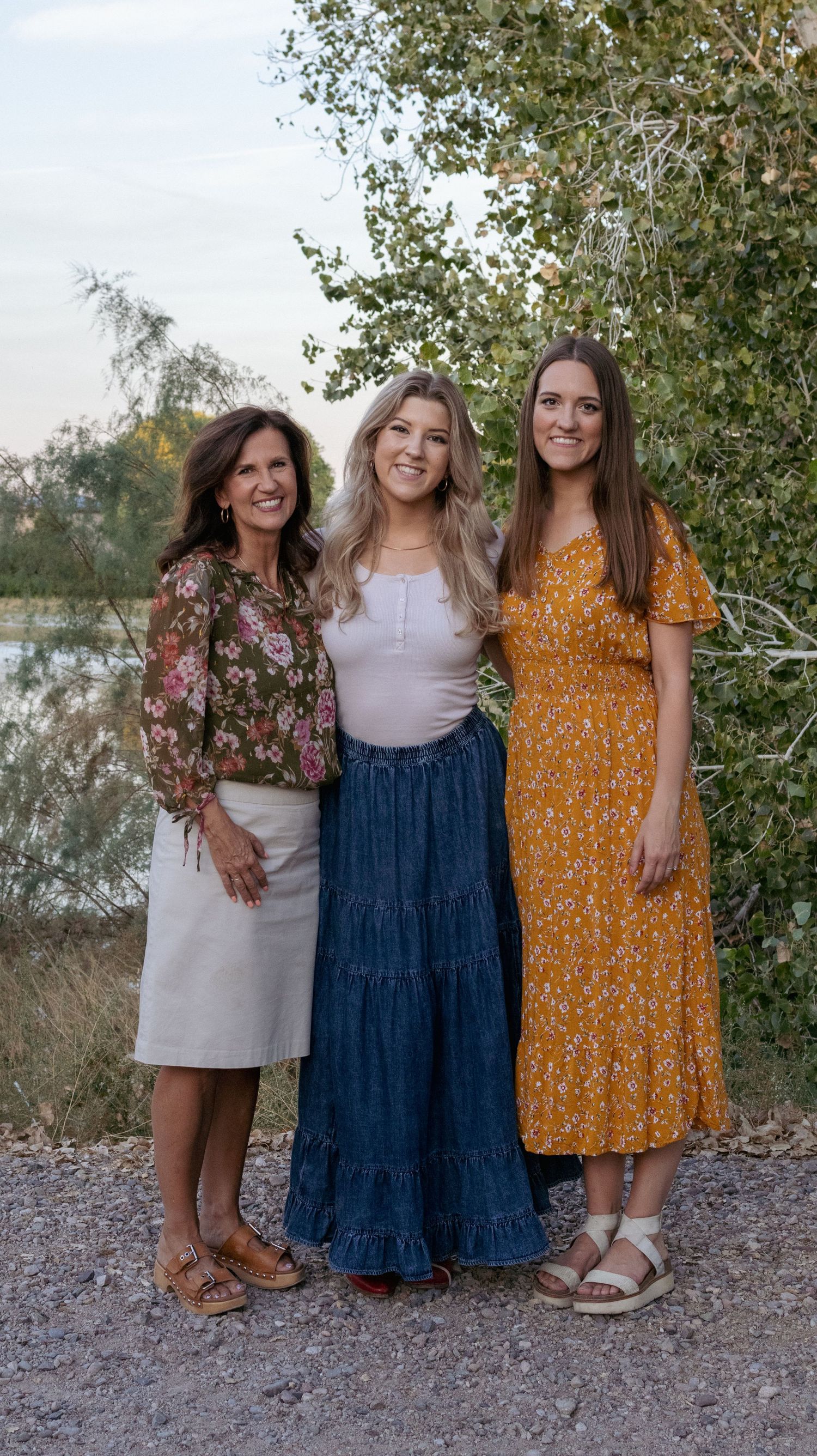 Three generations of women pose together on path wearing coordinated dresses in natural setting.