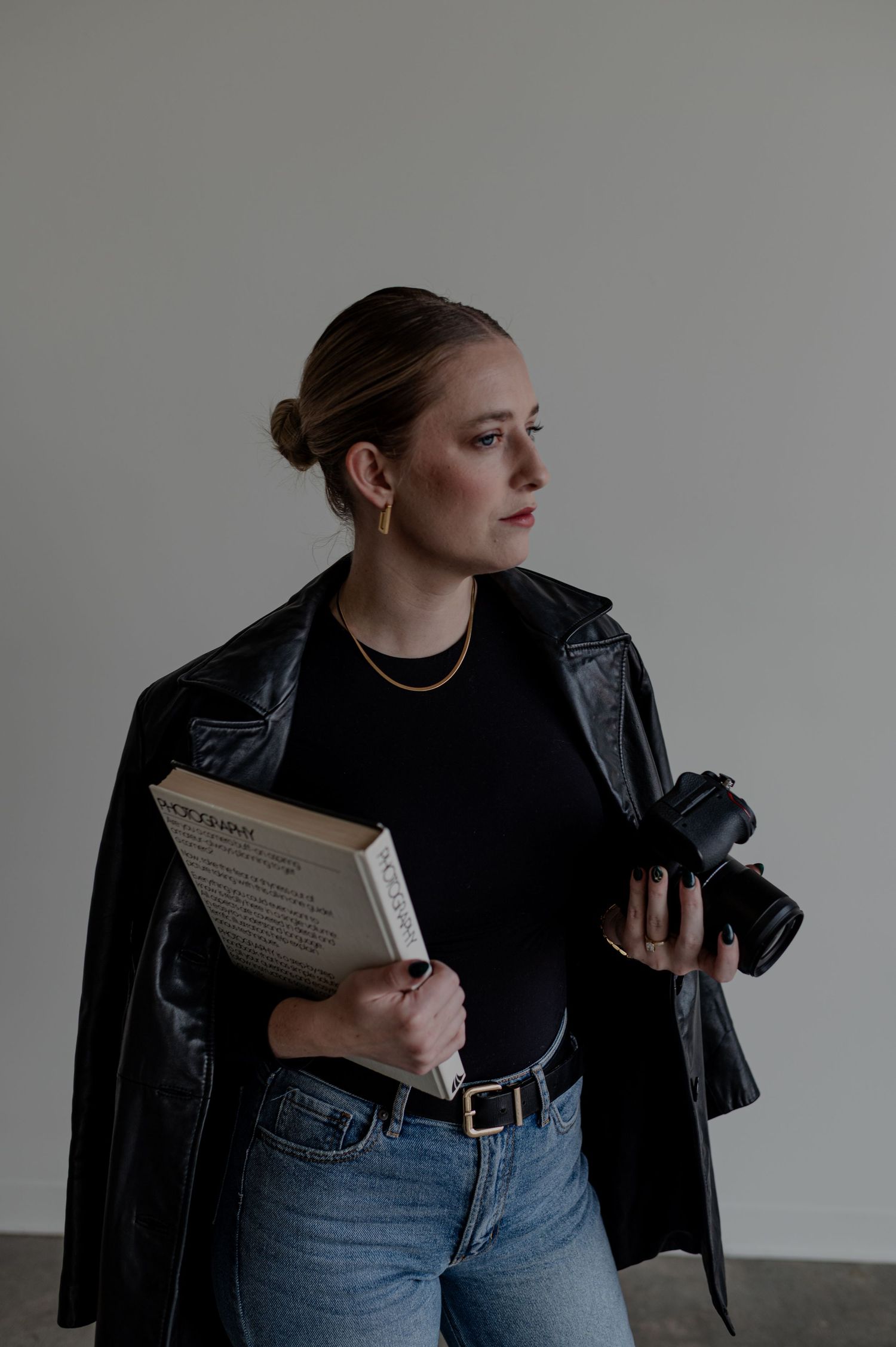 A photographer in casual attire holds a camera and book while wearing a leather jacket.
