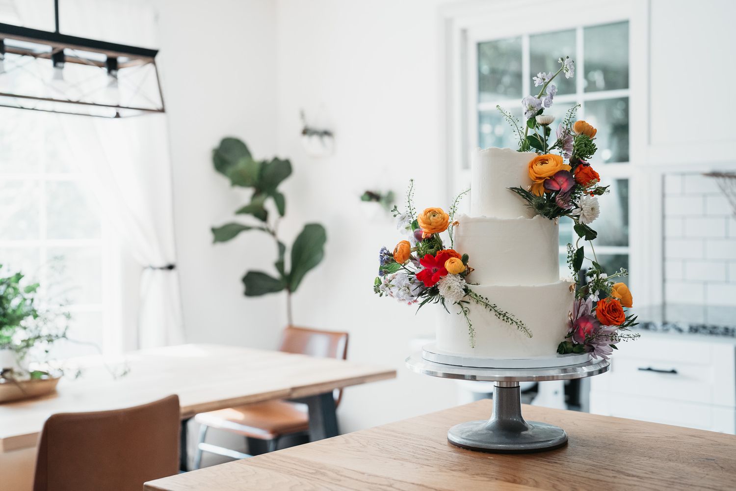 A three-tiered white wedding cake decorated with vibrant orange and red flowers sits on a black pedestal in a bright kitchen. Stow, Ohio