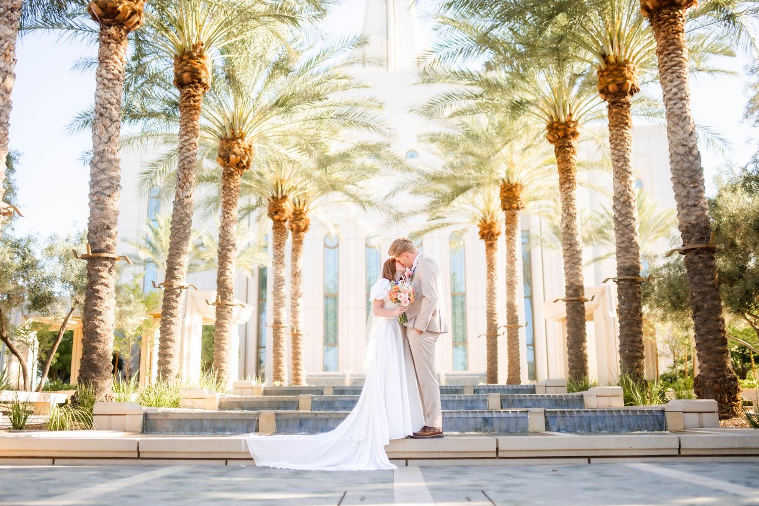 Bride and groom sharing romantic kiss under tall palm trees in sunlit outdoor wedding venue.
