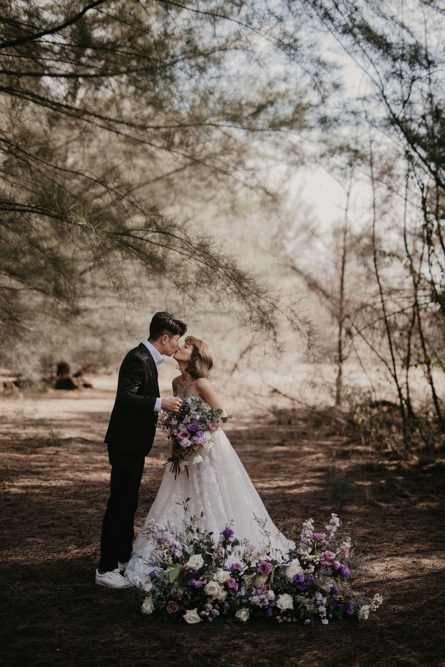Newlyweds share an intimate moment surrounded by scattered purple flowers in a wooded forest setting.
