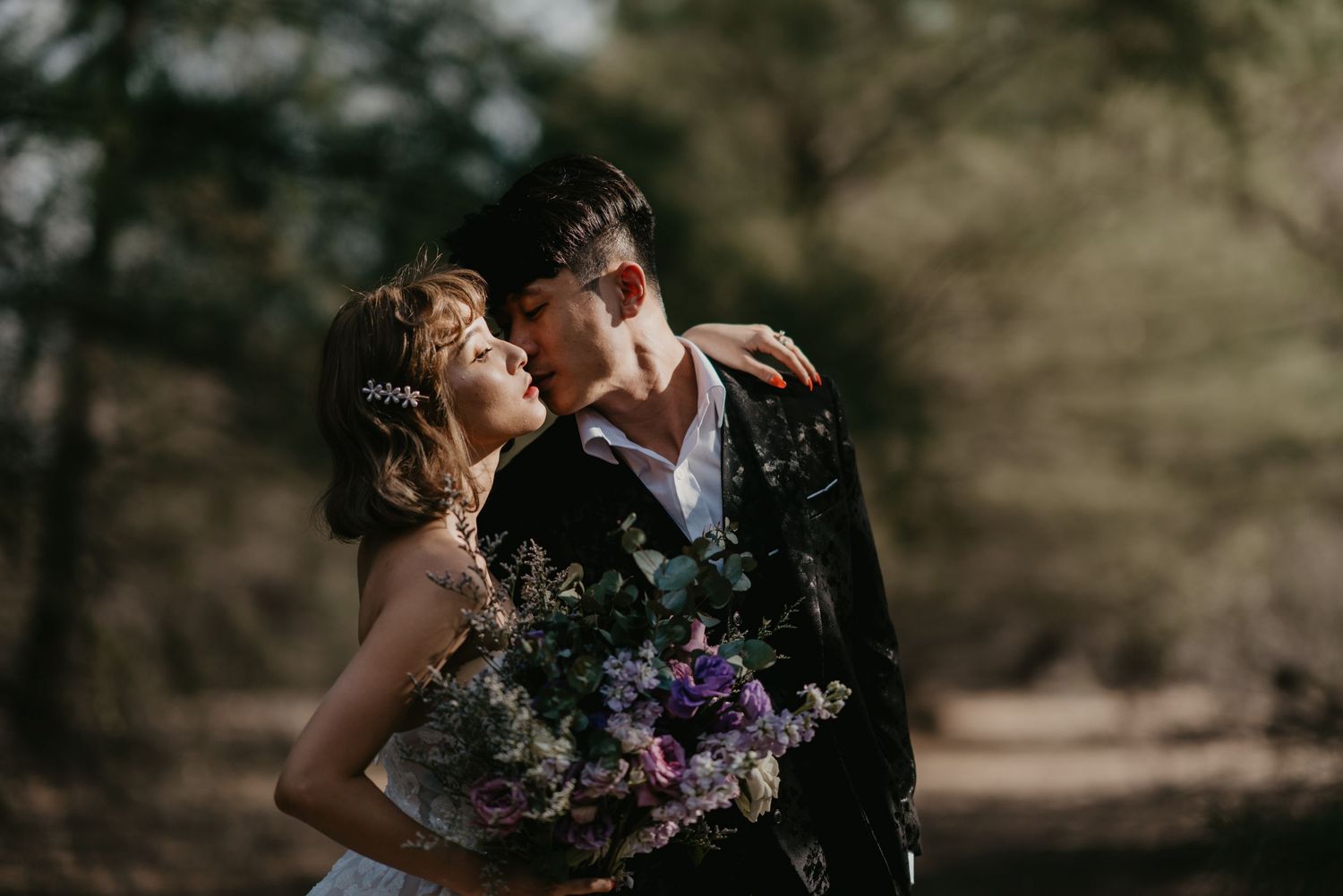 A tender moment between newlyweds captured in a moody forest setting with purple florals.