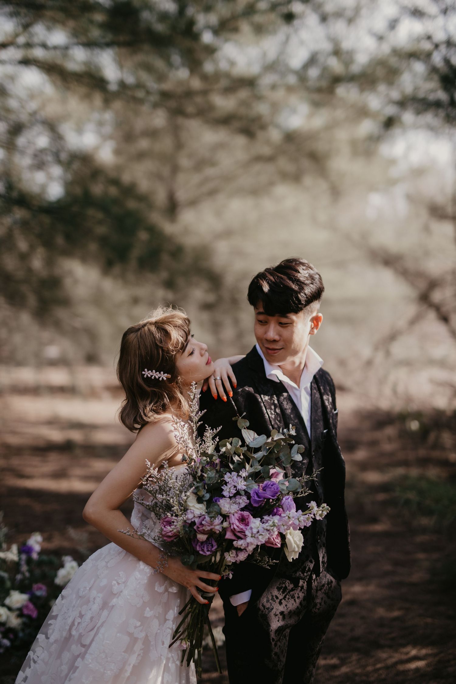 Wedding couple shares playful moments in a forest with scattered purple flower arrangements.