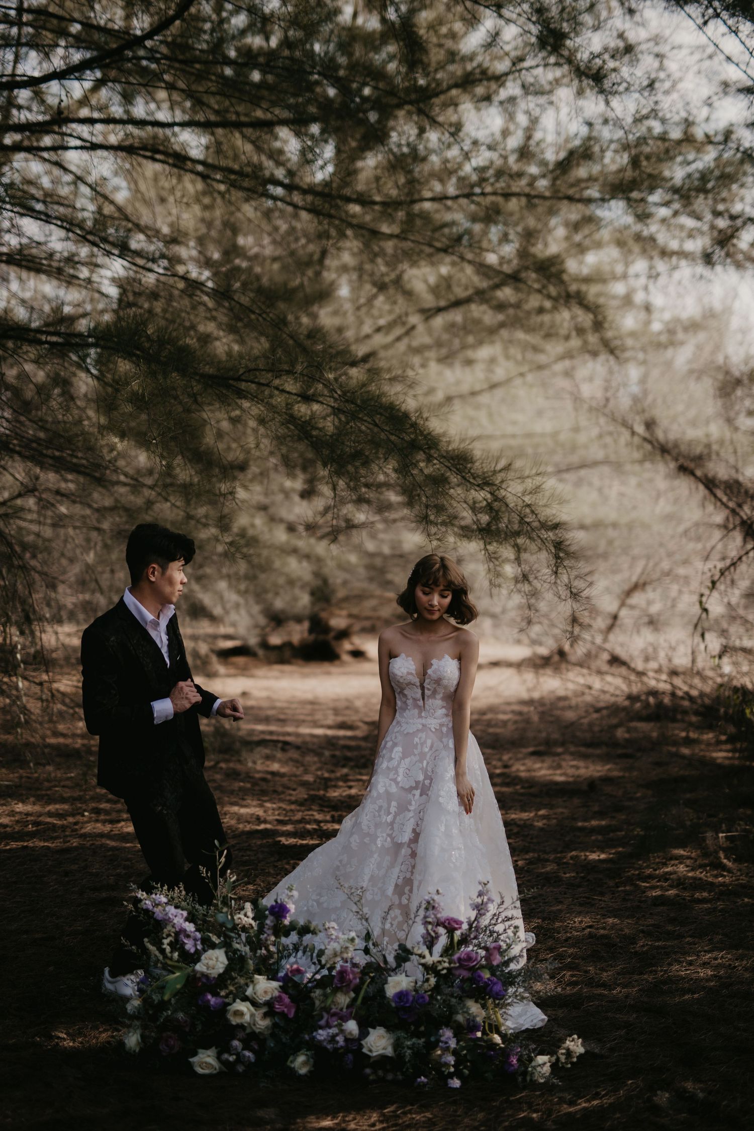 A couple poses with purple floral arrangements in a moody forest setting.