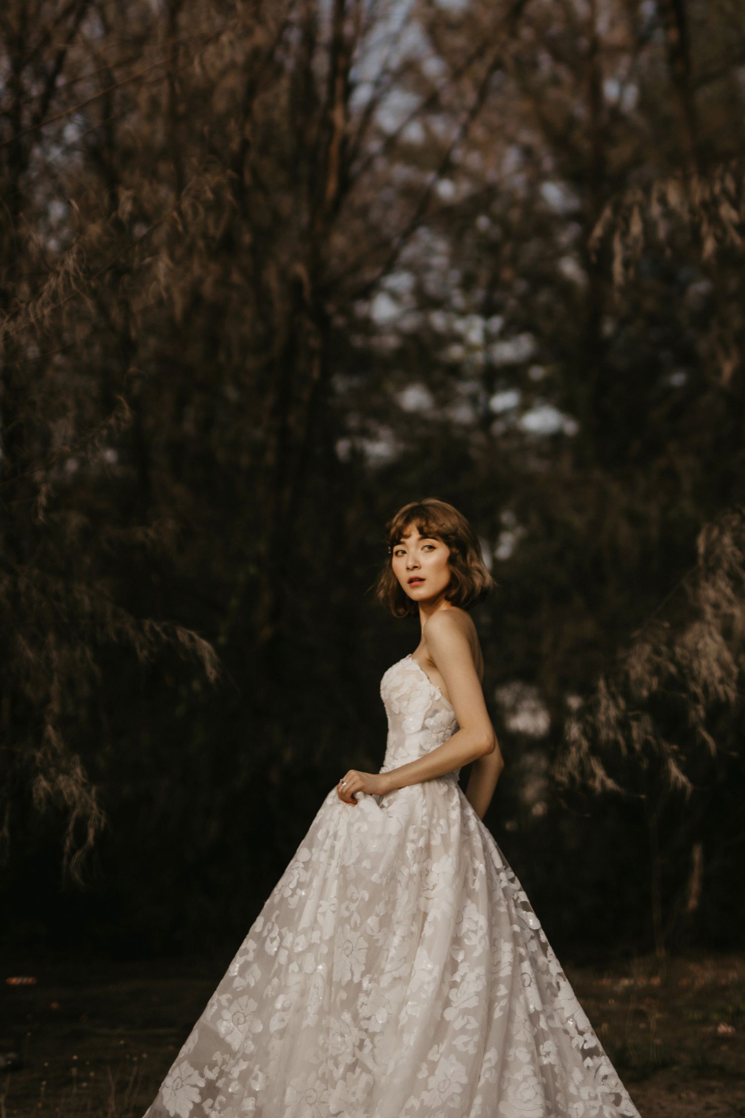 A bride in an elegant lace ballgown twirls in a moody forest setting.