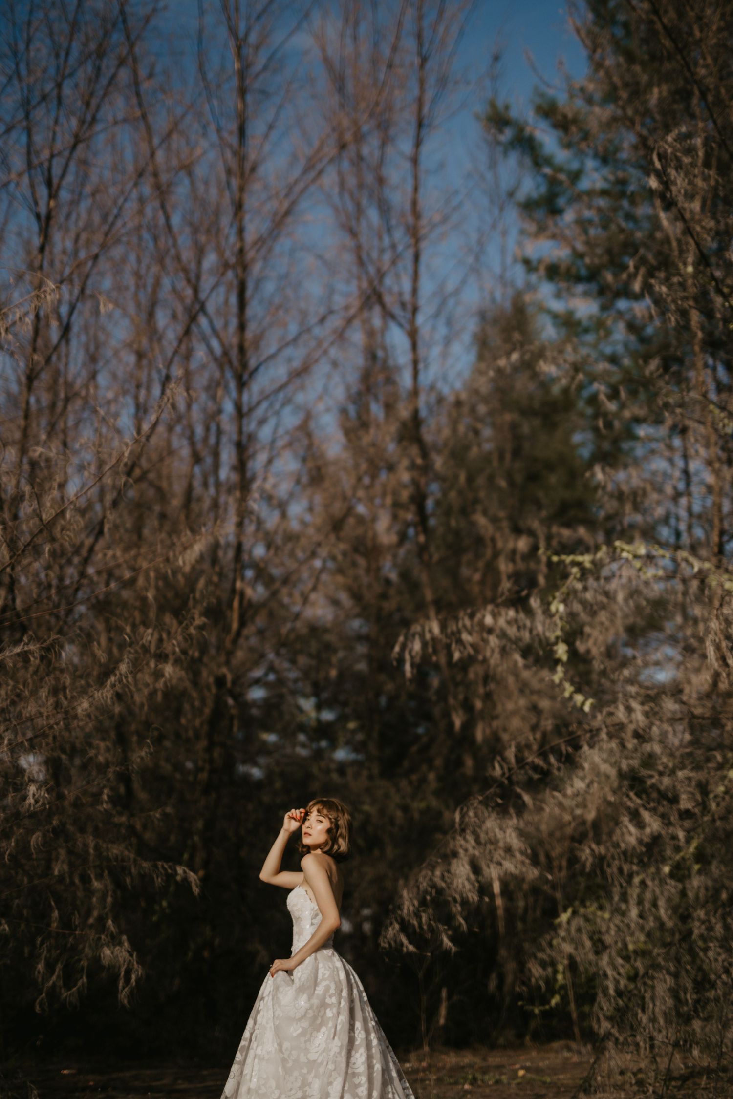 A bride in a flowing white dress poses against bare winter trees and blue sky.