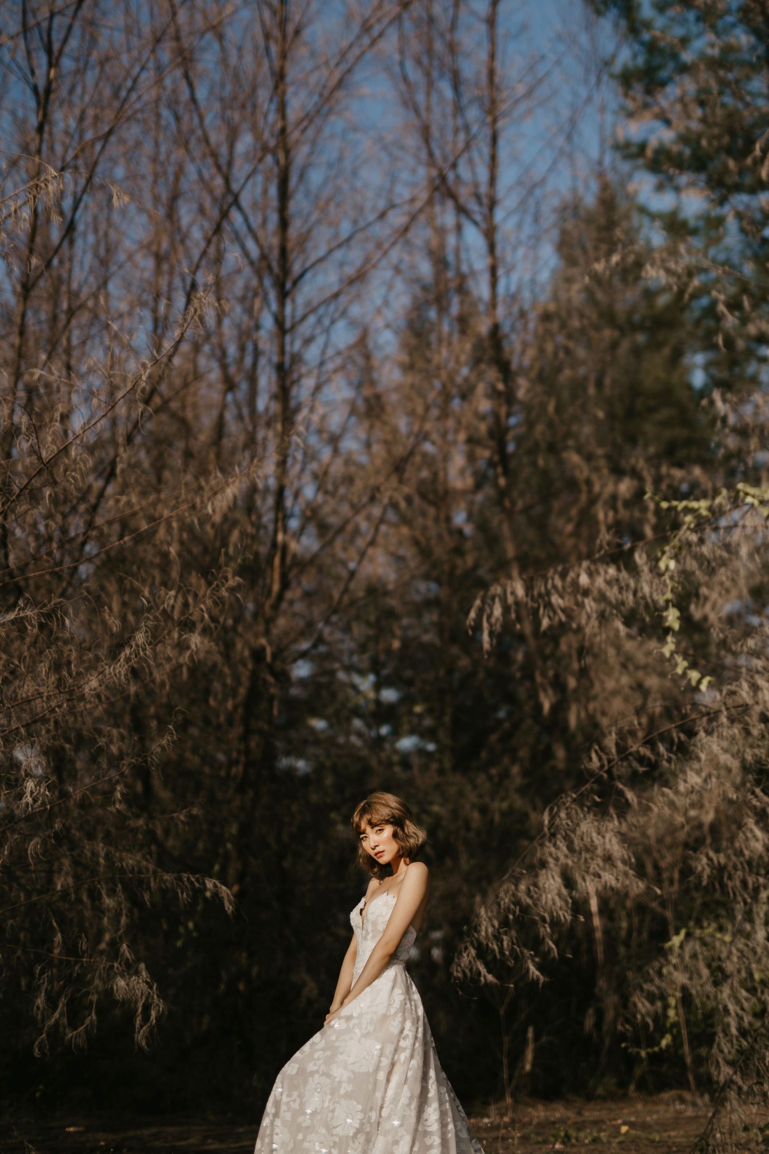 A bride in a detailed white gown poses elegantly among winter trees.