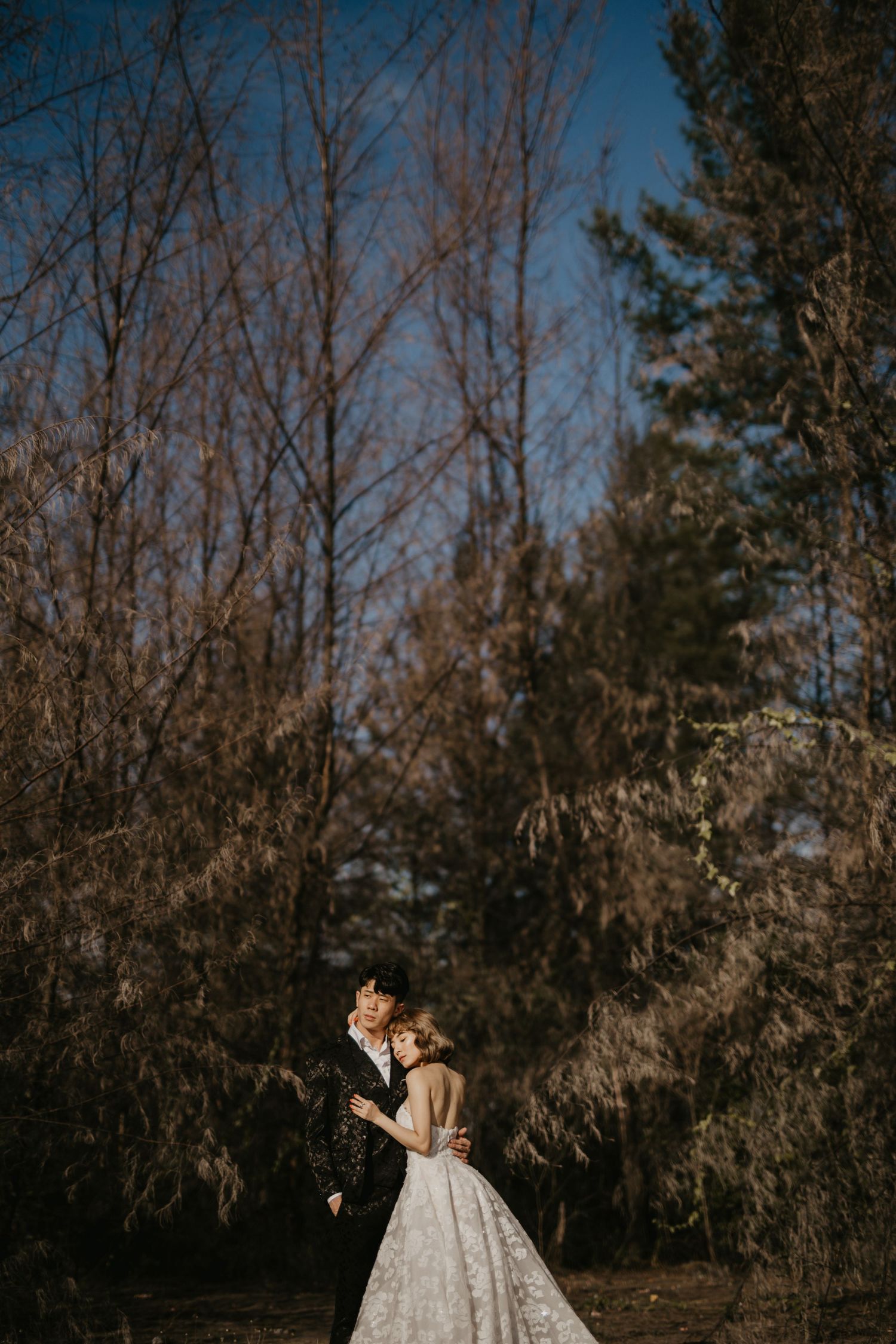 A wedding couple stands close together against a backdrop of tall trees.