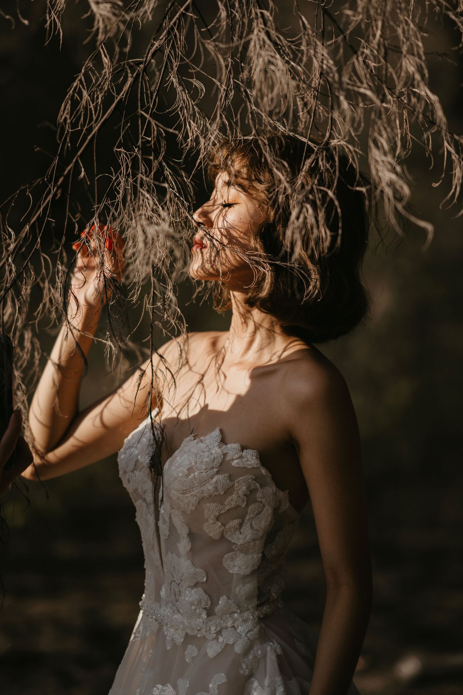 A bride in a lace dress poses artistically under hanging branches.
