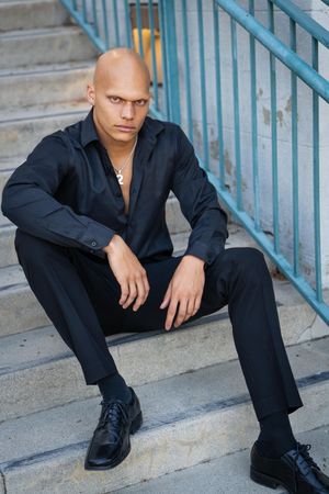 A person in black attire sits on concrete steps with a blue railing while posing for a series of artistic portraits.