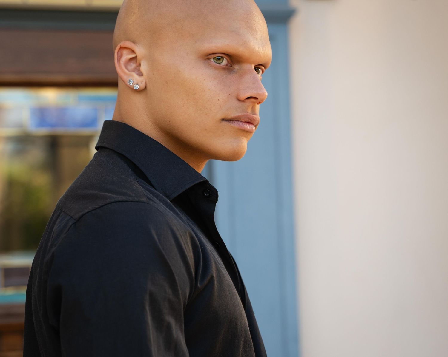 A person in a black collared shirt stands in profile against a light-colored wall with blurred background.