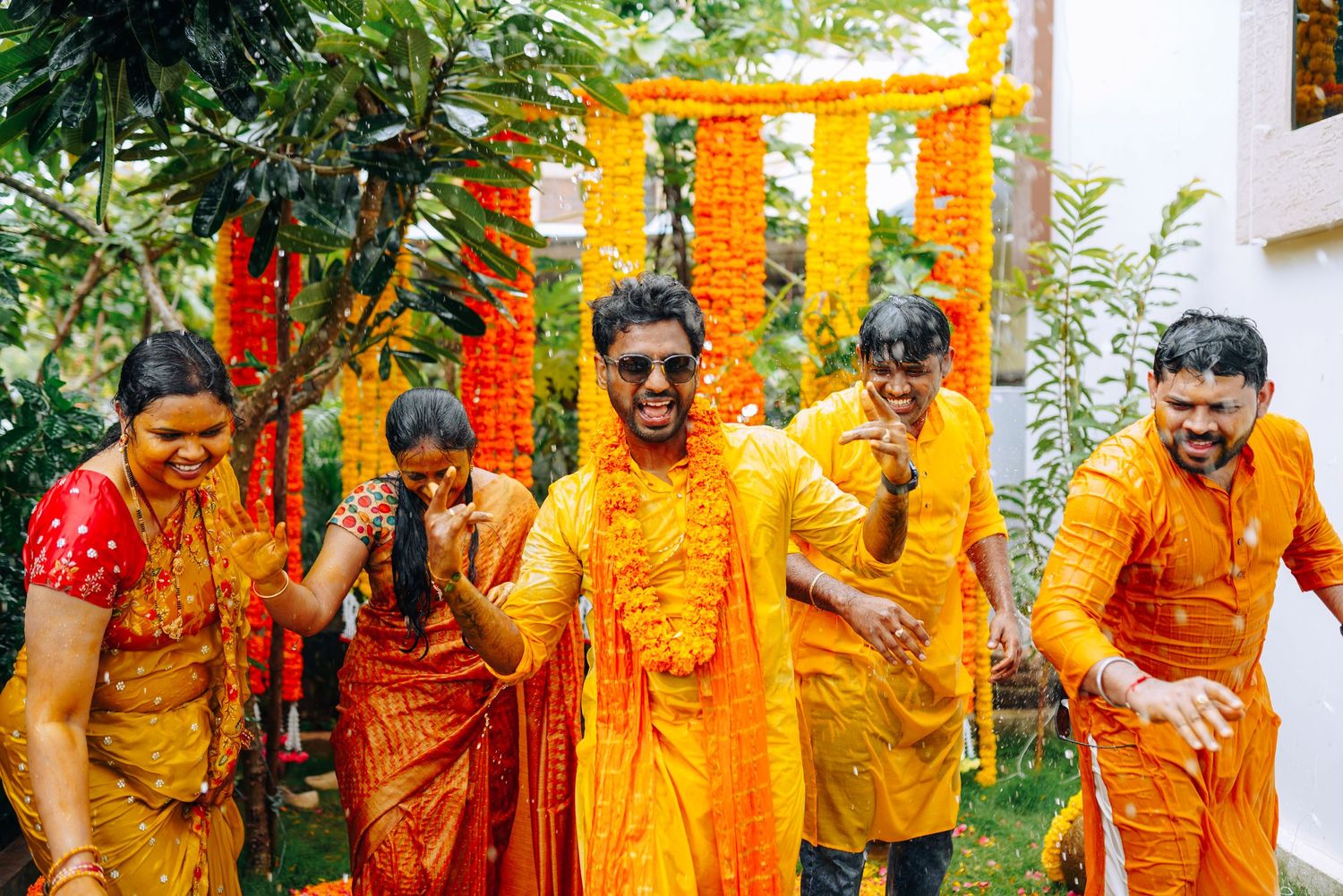 Group celebrating Holi festival covered in yellow powder under marigold decorations.