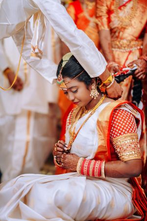Traditional South Indian bride in an ornate red and white saree with gold jewelry during wedding ceremony.