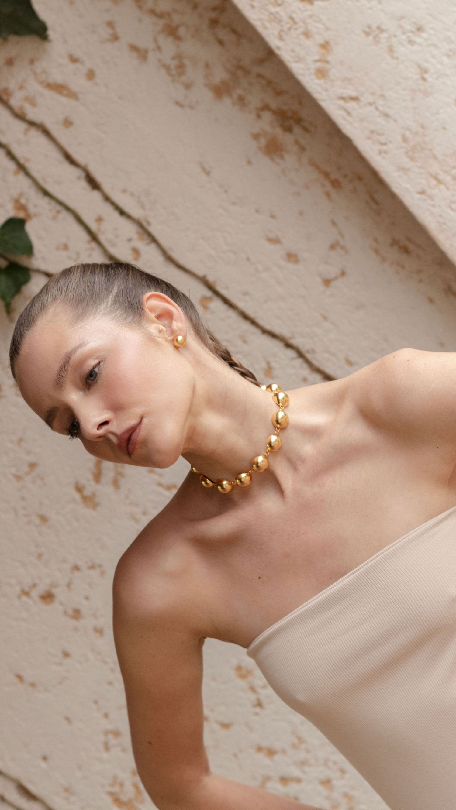 Fashion portrait featuring elegant gold ball necklace and strapless dress against textured wall.
