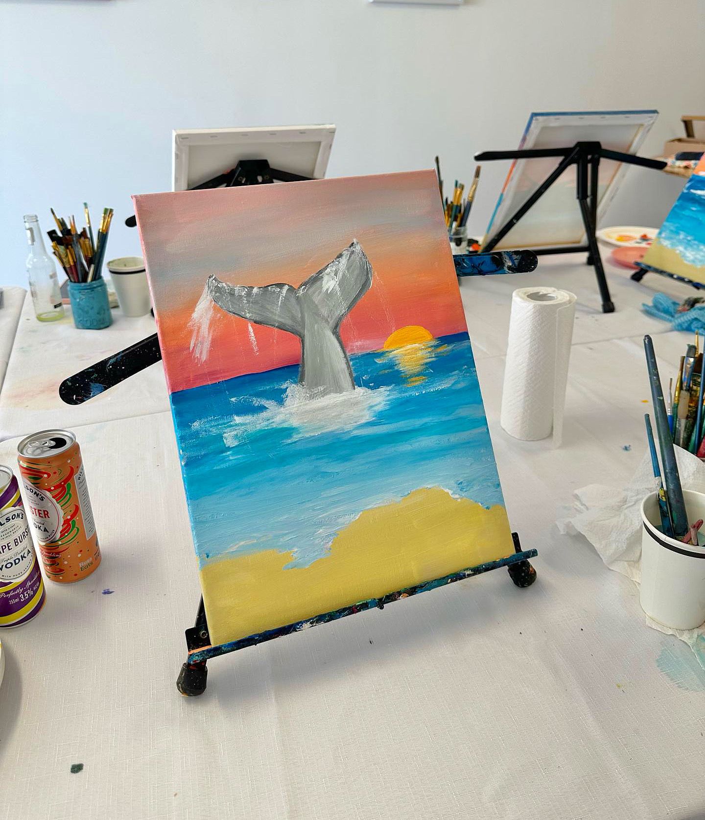 Painting of whale tail splashing in ocean during vibrant sunset on easel.
