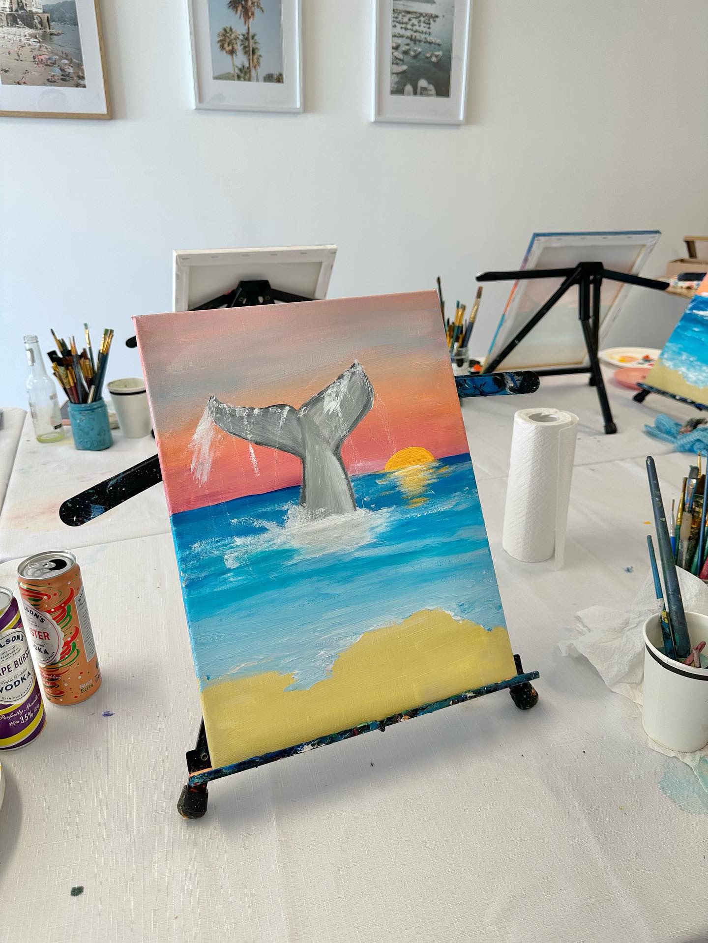Close-up of whale tail sunset painting with art supplies on white table.