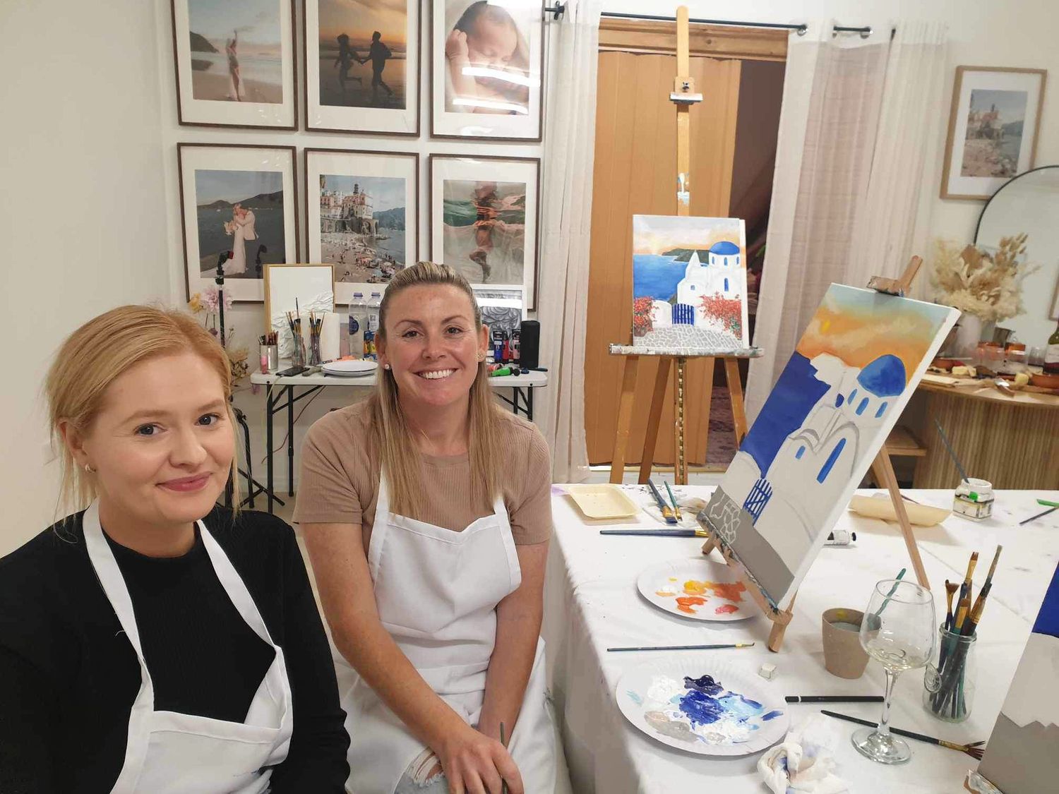 Two people wearing white aprons sit together in an art studio with paintings displayed on the wall behind them.