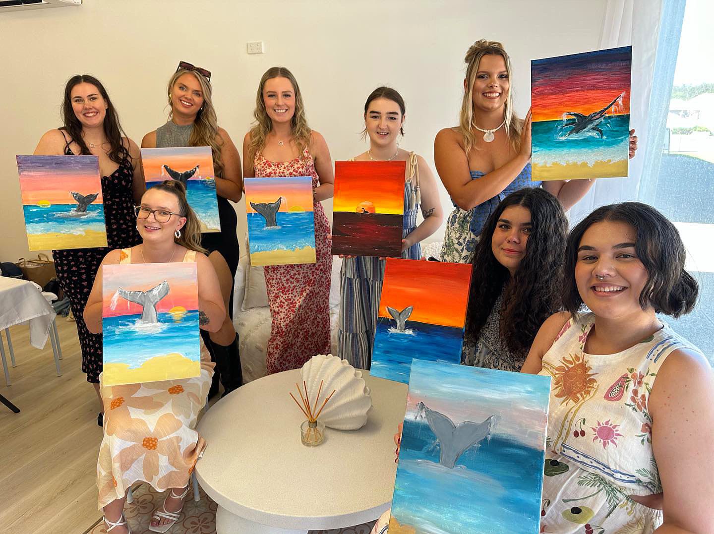 A group of art students proudly display their colorful beach sunset paintings during a painting class.