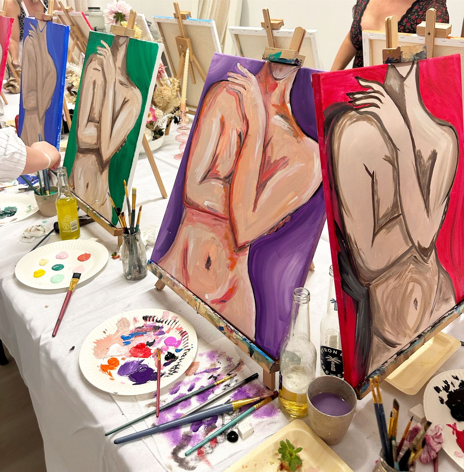 Artistic nude figure paintings displayed on easels with paint palettes and brushes on a white table.