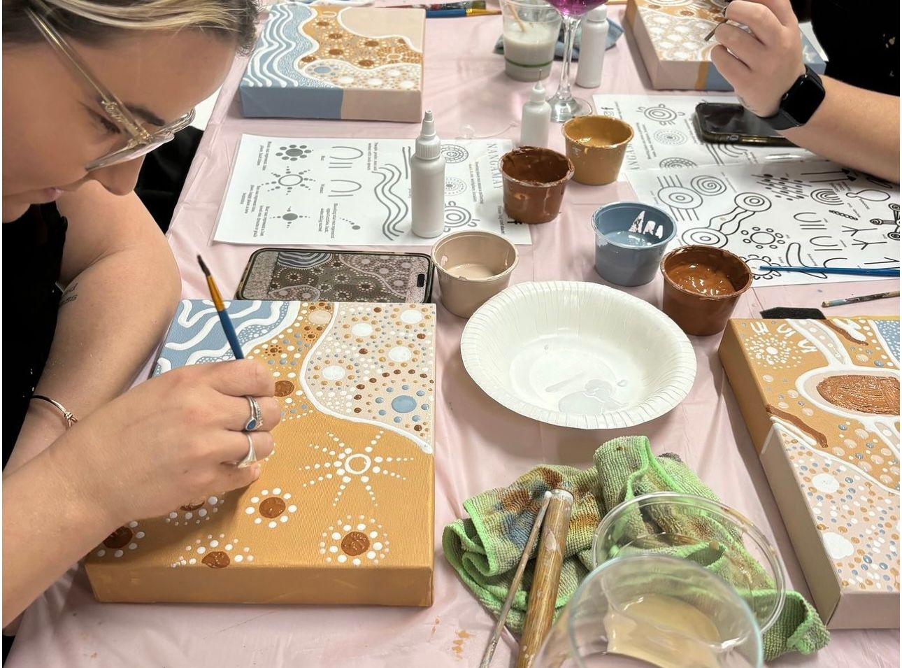 Students work on detailed mandala-style paintings with drinks and art supplies on the table.